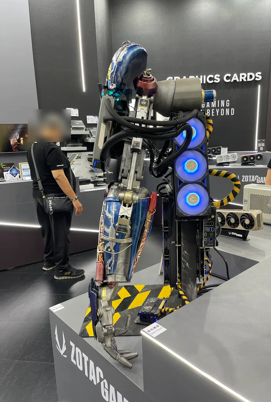 A mech-shaped gaming PC standing at the ZOTAC booth, with three blue glowing fans inside its body