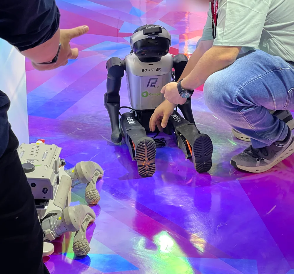 A small humanoid robot sitting on a colorful booth floor, with a staff member crouching beside it to adjust it