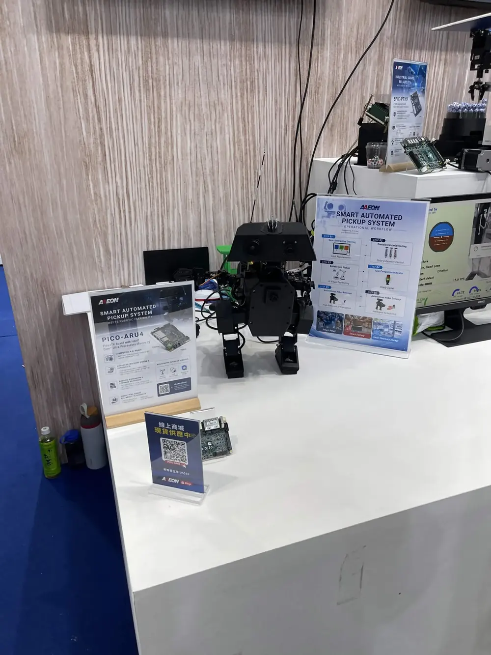 A small bipedal robot with two arms and cameras on a booth table, next to product signs