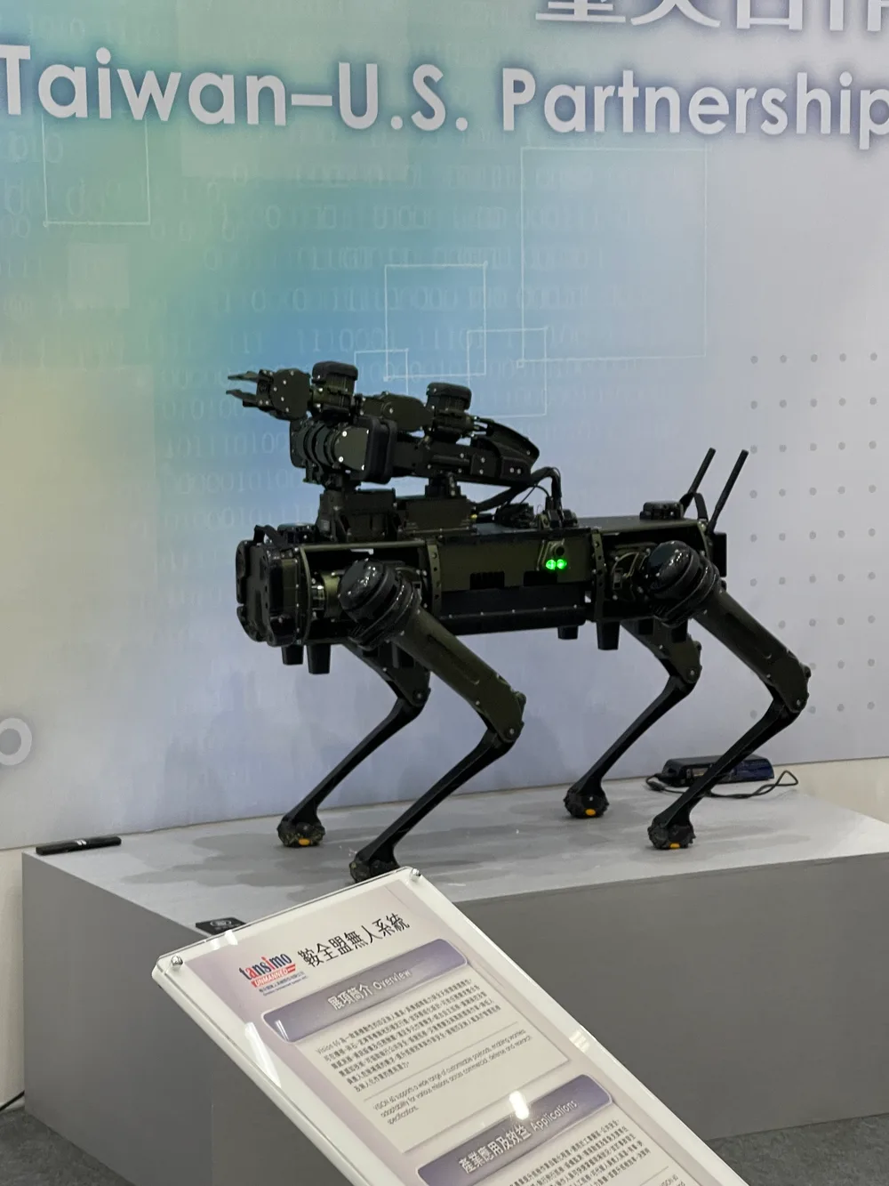 A black quadruped robot with a robot arm on its back, and a sign in front of the stand about unmanned systems