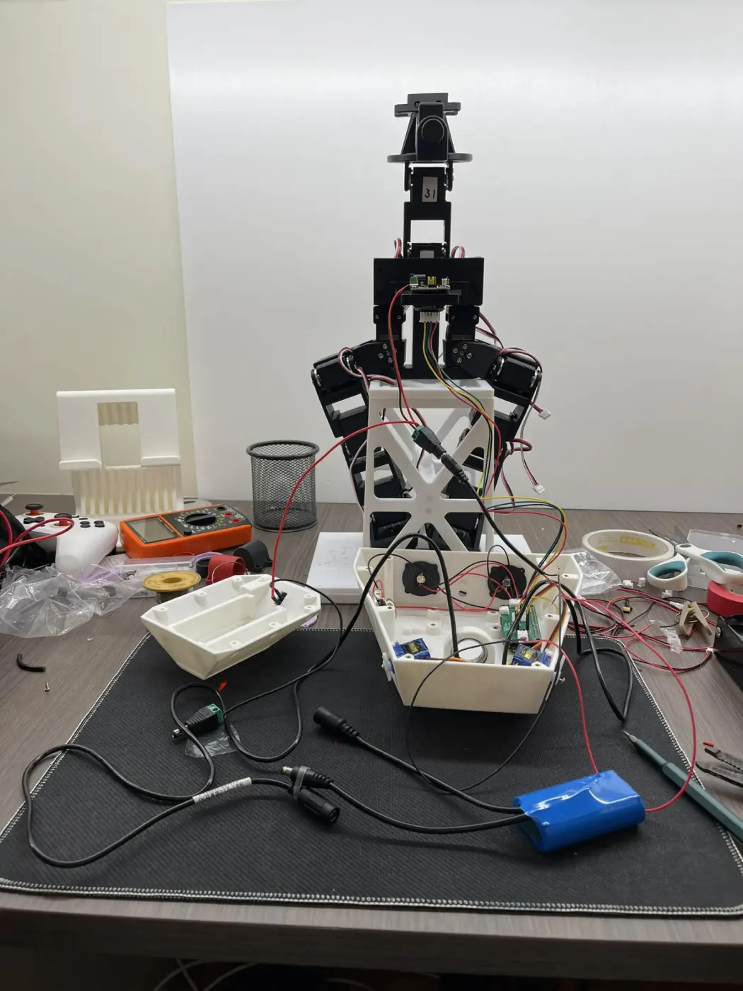 The robot on the desk with its head shell open, next to a blue 2S battery, DC cables, and a multimeter